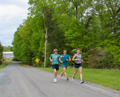 Three runners smiling and talking while jogging down a scenic, tree-lined paved road at a triathlon training camp.