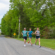 Three runners smiling and talking while jogging down a scenic, tree-lined paved road at a triathlon training camp.