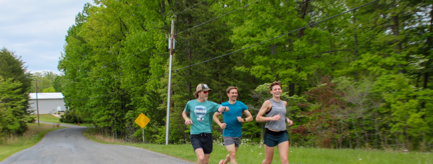 Three runners smiling and talking while jogging down a scenic, tree-lined paved road at a triathlon training camp.