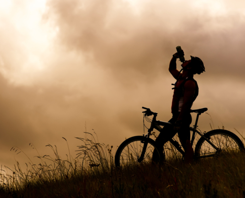 Silhouette of a cyclist drinking from a water bottle on a grassy hill under a dramatic sky, illustrating a personalized fueling and hydration strategy for endurance athletes.