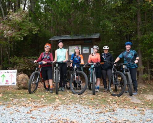 Group of six female mountain bikers standing with their bikes in front of a trail board at Le Tour de Femme.