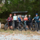 Group of six female mountain bikers standing with their bikes in front of a trail board at Le Tour de Femme.