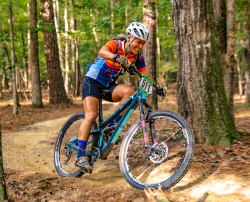 Coach Chris smiling as she rides a teal full-suspension bike through a wooded trail during a Roadless Races event.