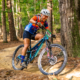 Coach Chris smiling as she rides a teal full-suspension bike through a wooded trail during a Roadless Races event.