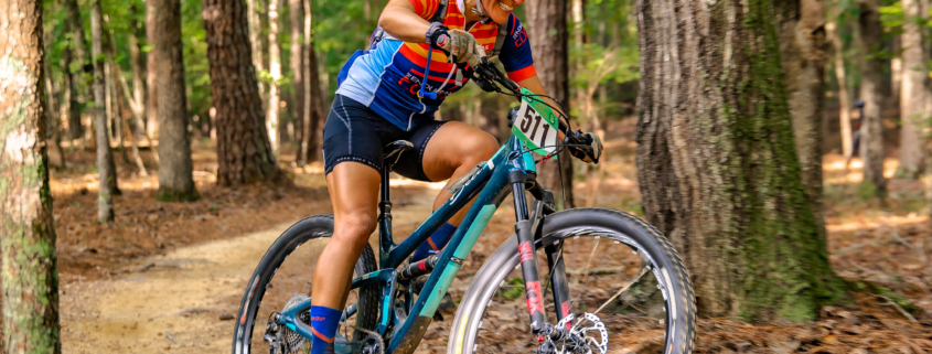 Coach Chris smiling as she rides a teal full-suspension bike through a wooded trail during a Roadless Races event.