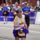 A determined elderly woman with grey, curly hair smiles broadly while running the Boston Marathon, wearing a "New England 65+ Runners Club" shirt and race bib number 32012. Her participation exemplifies lasting motivation in endurance sports.