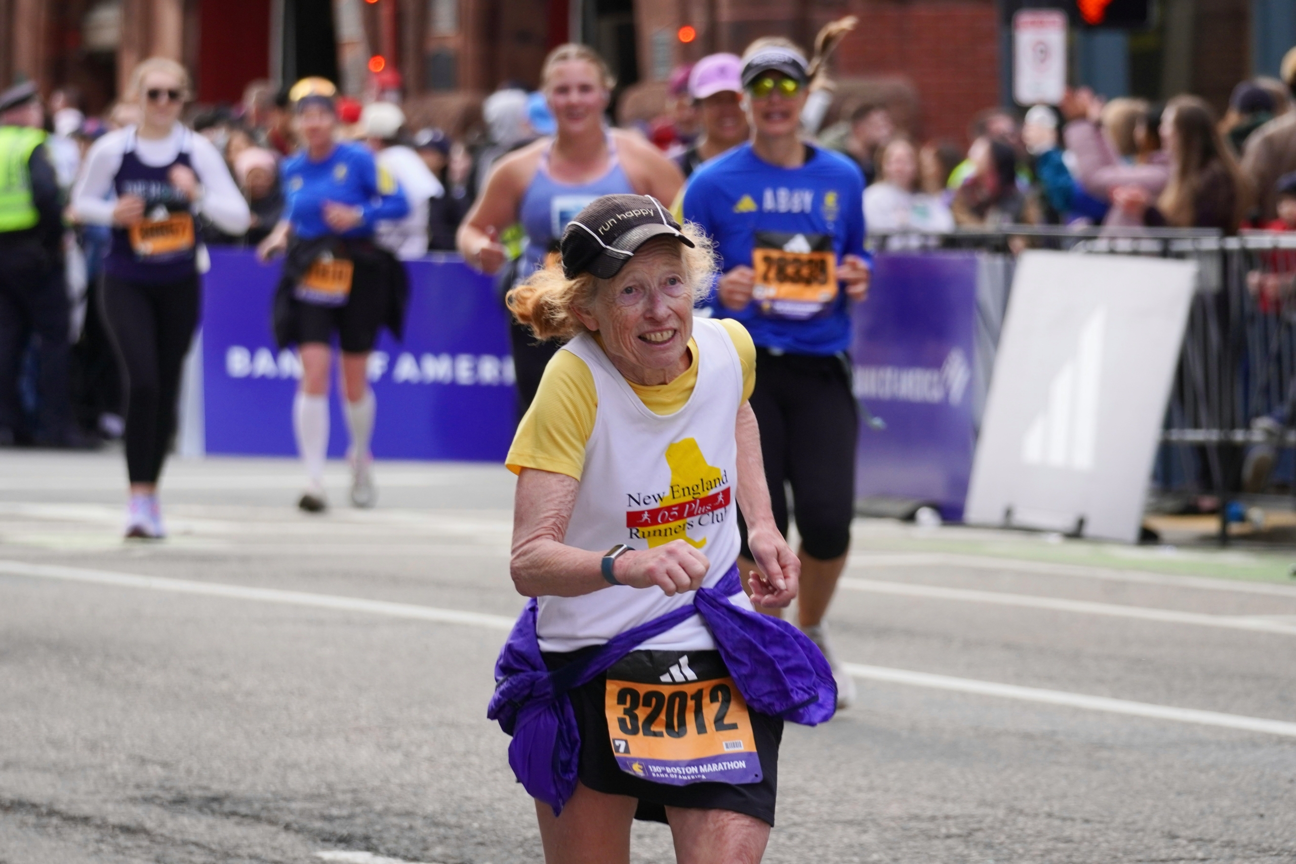 A determined elderly woman with grey, curly hair smiles broadly while running the Boston Marathon, wearing a "New England 65+ Runners Club" shirt and race bib number 32012. Her participation exemplifies lasting motivation in endurance sports.
