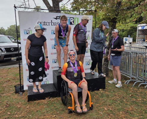 Kris Bonot smiling on an adaptive triathlon podium in an orange wheelchair surrounded by fellow competitors at the Outer Banks Tri event.