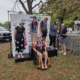Kris Bonot smiling on an adaptive triathlon podium in an orange wheelchair surrounded by fellow competitors at the Outer Banks Tri event.