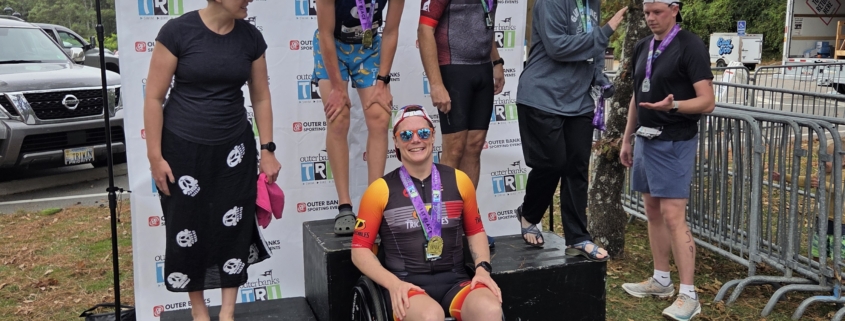 Kris Bonot smiling on an adaptive triathlon podium in an orange wheelchair surrounded by fellow competitors at the Outer Banks Tri event.