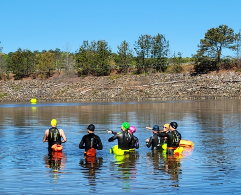 A group of swimmers wearing wetsuits, bright caps, and safety buoys standing in shallow lake water during an open water swimming for beginners session.